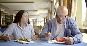 Husband bald and glasses and dark-haired middle-aged wife, sitting in a cafe, eat, communicate. In the hands of they