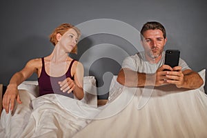 Husband or boyfriend using mobile phone in bed and suspicious frustrated wife or girlfriend feeling upset suspecting betrayal and