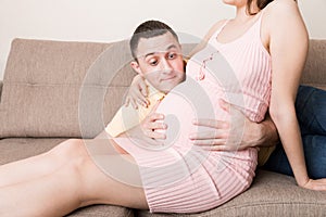 Husband listening to his pregnant wife`s belly on sofa at home