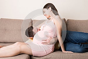 Husband listening to his pregnant wife`s belly on sofa at home