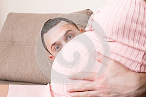 Husband listening to his pregnant wife`s belly on sofa at home