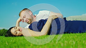 Husband with a pregnant wife is lying on the green grass. Against the background of the blue sky