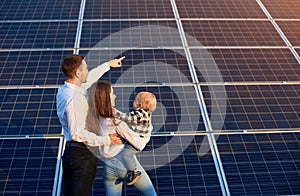 Husband shows his family the solar panels on the background. Blond child in arms of wife.