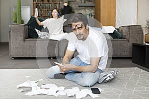 Husband sitting on the floor with many bills and calculating expenses of his wife who sitting on the couch