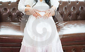 Husband uses hand to touch belly of pregnant wife in white dress, sitting on sofa brown leather ,concept pregnancy birth of new