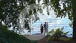 Husband and wife and child stand by water in summer. Stock footage. Beautiful view through branches of birch on family