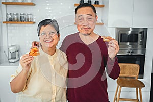 Husband and wife happily holding pizza in the kitchen and looking at the camera