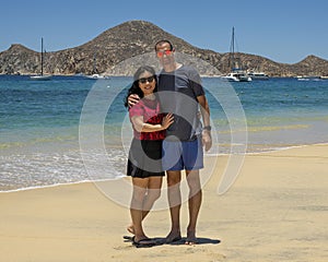 Husband and wife standing in the sand of Medano Beach with the Sea of Cortez and moored boats in the background in Cabo San Lucas.