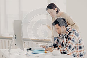 Husband and wife work on paper report together, check information, sit in front of computer, drinks coffee, dressed in casual wear
