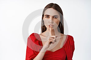 Hush dont speak. Young beautiful woman in red dress tell to keep quiet, taboo gesture, press finger to lips has secret