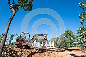 Hydraulic excavator is digging soil at a construction site, surrounded by lot of trees, under clear sky. Clawer backhoe parked