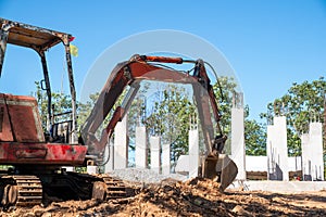 Hydraulic excavator is digging soil at a construction site, surrounded by lot of trees, under clear sky. Clawer backhoe parked