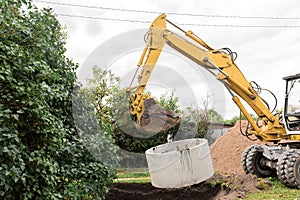 Hydraulic piston system of the excavator with a bucket, lowering into the pit on steel cables concrete sewer ring. Construction or