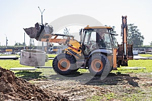 Hydraulic piston system of the excavator with a bucket, lowering into the pit on steel cables concrete sewer ring. Construction or