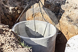 Hydraulic piston system of the excavator with a bucket, lowering into the pit on steel cables concrete sewer ring. Construction or