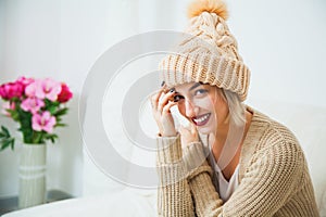 Hygge, comfort and home concept. Young woman in warm beige hand knitted hat at home. Pretty lady in big wool cap, smiling happily