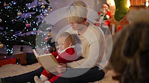 Hyperactive baby girl in Santa costume gesturing as woman reading book for toddler. Carefree Caucasian kid resting at
