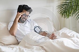 Hypertension. Indian Man Checking Blood Pressure With Upper Arm Monitor In Bed