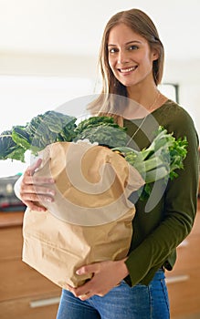 I only buy the freshest produce. Portrait of a smiling young woman standing in her kitchen carrying a paper bag full of