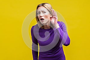 I can`t hear! Portrait of curious attentive woman keeping hand near ear. indoor studio shot isolated on yellow background