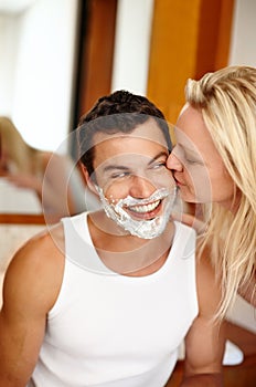 I cant keep my hands off of him. A handsome man with shaving cream on his face smiling while his girlfriend kisses his