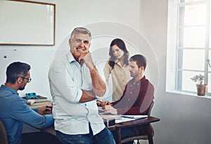 I couldnt be happier with my team. Portrait of a mature man sitting on a desk in the office which his colleagues are