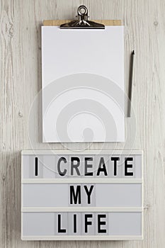 `I create my life` words on a lightbox, clipboard with blank sheet of paper on a white wooden surface, top view. Overhead, from