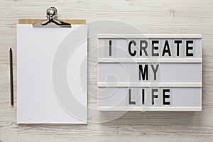 `I create my life` words on a lightbox, clipboard with blank sheet of paper on a white wooden surface, top view. Overhead, from