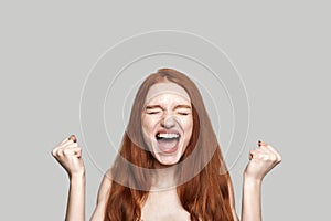 I did it Portrait of happy young redhead woman screaming and keeping fists clenched while standing against grey