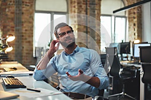 I don`t hear you. Portrait of young bearded man in eyeglasses and headphones talking with client while sitting in the