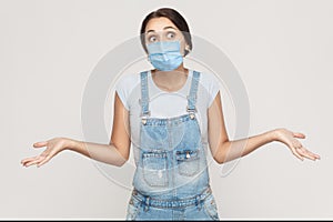 I don`t know. Portrait of thoughtful young brunette woman with surgical medical mask in denim overalls standing raised arms and