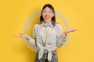 I don't know. Puzzled young Asian woman shrugging shoulders and smiling at camera on yellow studio background