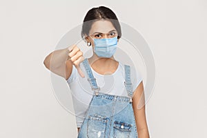 I don`t like it. Portrait of dissatisfied young brunette woman with surgical medical mask in denim overalls standing thumbs down