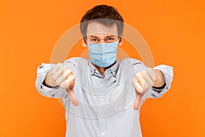 I don`t like it. Portrait of sad young worker man with surgical medical mask standing thumbs down and looking at camera with