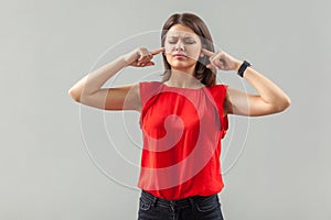 I don`t want to hear you. Portrait of confused brunette young woman in red shirt standing with closed eyes and putting her finger