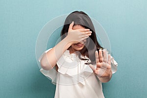 I don`t want to see this. Portrait of scared girl with black straight hair in white dress standing, covering her eyes and showing