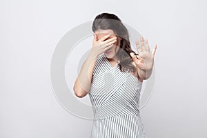 I don`t want to see this. young brunette woman with striped dress standing covering her face and showing stop gesture and try to