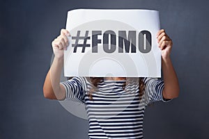 I feel so excluded. Studio shot of a young woman holding a sign with FOMO printed on it against a gray background.