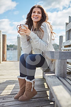 I feel fall in the air. a beautiful young woman enjoying a warm beverage while relaxing on a bench at a lake.