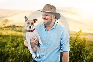 I have a buddy who helps me on the farm. a farmer holding his dog in a vineyard.