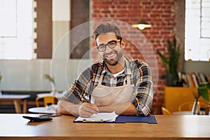 I keep my business running smoothly. Portrait of a happy young business owner doing admin at a table in his coffee shop.