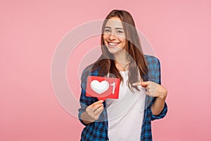 I like this content! Charming positive girl in checkered shirt smiling playfully and holding heart icon