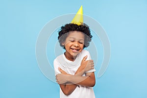 I love and congratulate myself. Portrait of amazing lovely kid with funny party cone embracing himself and smiling from pleasure