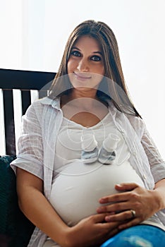 I love my baby bump. Portrait of a happy pregnant woman sitting in a chair at home with a pair of baby shoes on her