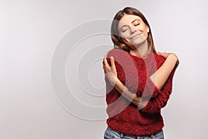 I love myself! Portrait of selfish girl in shaggy sweater embracing herself, closed eyes and smiling with pleasure