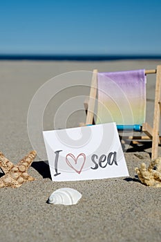 I LOVE SEA text on paper greeting card on background of beach chair lounge starfish summer vacation decor. Sandy beach