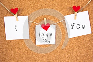 I love you handwriting lettering on 3 white papers pinned with 2 big red heart pegs and one small red heart pegs on cork board