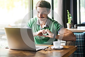 I love you. Young happy romantic businessman in green t-shirt sitting and looking ot laptop screen on video call with love gesture