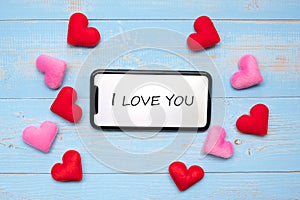 I LOVEYOU word on white touchscreen display of black smart phone with red and pink hearts shape decoration on blue wooden table photo