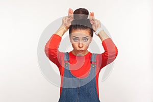 I`m a cow! Portrait of angry girl with hair bun in denim overalls standing with bull horns, antler gesture on her head